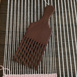 May include: A dark brown wooden comb with a handle, used for weaving. The comb is lying on a white warp thread on a loom. The loom has a pink, green, and white woven tapestry.