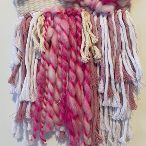 Op de afbeelding: Een geweven wandkleed in roze en wit met lange franjes. De franjes zijn gemaakt van verschillende kleuren garen, waaronder roze, wit en cr&egrave;me.