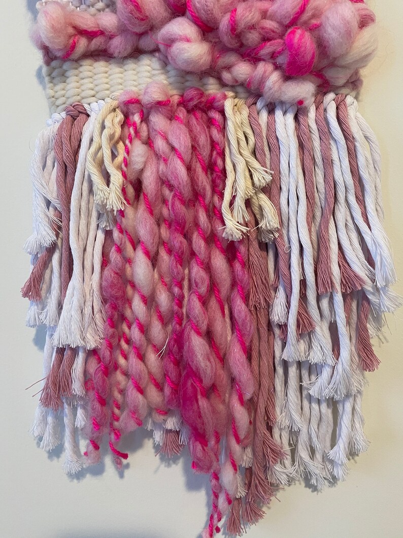 Op de afbeelding: Een geweven wandkleed in roze en wit met lange franjes. De franjes zijn gemaakt van verschillende kleuren garen, waaronder roze, wit en cr&egrave;me.