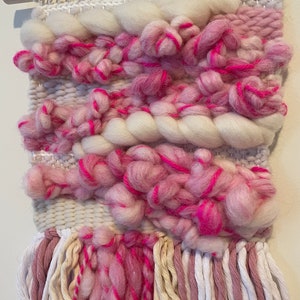Op de afbeelding: Een geweven wandkleed in roze en wit met franjes. Het kleed is gemaakt met verschillende texturen en kleuren, waaronder roze, wit en cr&egrave;me.