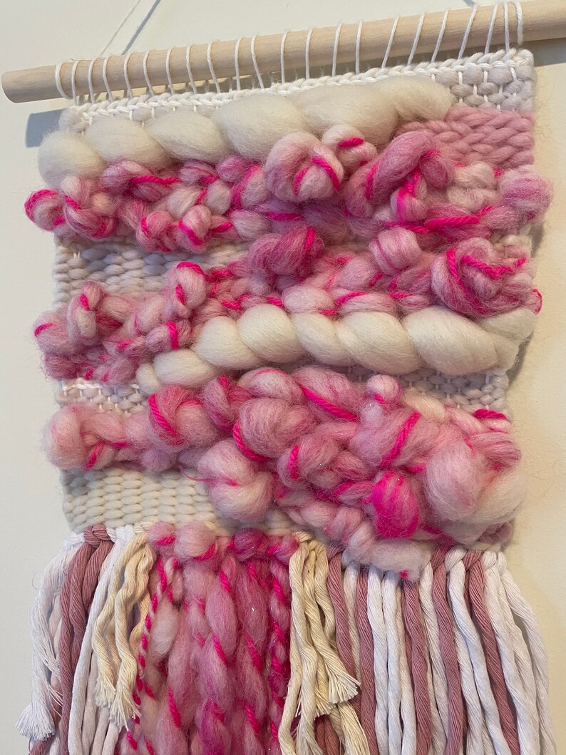 Op de afbeelding: Een geweven wandkleed in roze en wit met franjes. Het kleed is gemaakt met verschillende texturen en kleuren, waaronder roze, wit en cr&egrave;me.