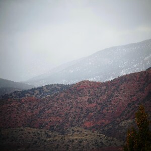 May include: A scenic landscape featuring layered mountain ranges under a cloudy sky. The foreground shows reddish-brown hills dotted with dark green vegetation. The distant mountains fade into a misty, light gray atmosphere.
