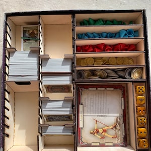 May include: A wooden box with compartments for organizing a board game. The box contains cards, dice, tokens, and a character card with the text "AMAZONE - Elfgriechen".