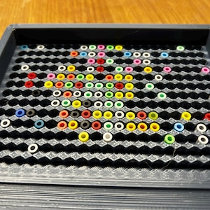 Könnte beinhalten: Eine schwarze Kunststoff-Pegboard-Schale gefüllt mit bunten Stiften. Die Stifte sind in einem Muster angeordnet, mit Farben wie Rot, Gelb, Grün, Blau, Rosa und Weiß. Die Schale hat ein gitterartiges Design.