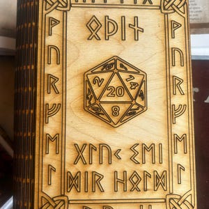 Könnte beinhalten: Eine holzfarbene, buchförmige Box mit gelaserten Runen und einem 20-seitigen Würfeldesign. Die Box besteht aus hellem Holz mit dunkelbraunen geätzten Details. Der Text "MI+YIX", "Xrn<EMI", "MIR H&IM" und "B&H" ist sichtbar.