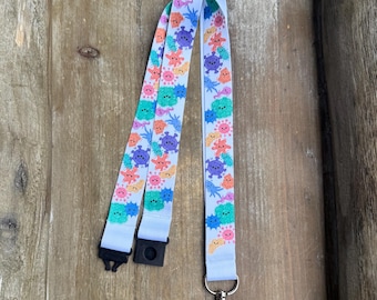 May include: A white lanyard with a colorful pattern of cartoon microbes. The lanyard has a black plastic clip and a silver metal clasp. The design features various microbes in shades of blue, green, orange, and purple.