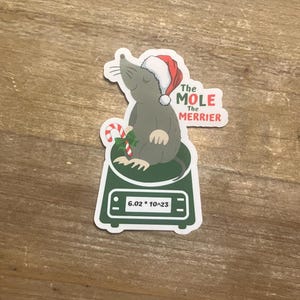 May include: A gray mole wearing a red Santa hat sits on a green scale. The scale reads "6.02 * 10^23". The text "The Mole The Merrier" is written above the mole.
