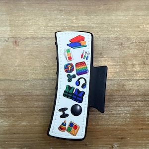May include: A black hair claw clip featuring a white rectangular center panel decorated with colorful miniature icons including building blocks, a pop-it toy, headphones, life vests, a fidget spinner, a stacking ring toy, and various other small shapes.