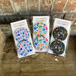May include: Three sets of car coasters with different science-themed designs. The first set features beakers and test tubes, the second set features colorful microbes, and the third set features a black and white doodle design with beakers, test tubes, and other science-related items. Each set has the text "Car Coasters" and "Handmade with love" on the packaging.