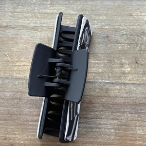 May include: A black and white patterned hair claw clip. The clip is made of plastic and has a spring mechanism. The claw clip is designed to hold hair in place. The clip is a hair accessory.