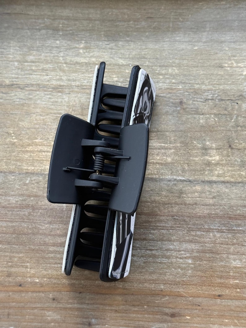 May include: A black and white patterned hair claw clip. The clip is made of plastic and has a spring mechanism. The claw clip is designed to hold hair in place. The clip is a hair accessory.