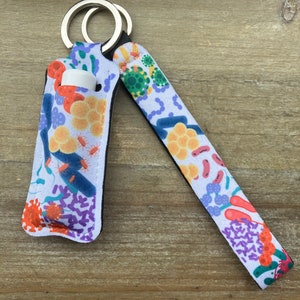 May include: A white lip balm holder with a colorful pattern of bacteria and viruses. The holder has a silver key ring and a black strap.