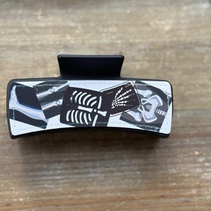 May include: Black hair claw clip featuring a design of x-ray images of bones and a skull. The clip is rectangular with a curved top and a matte finish. The x-ray images are in black and white.