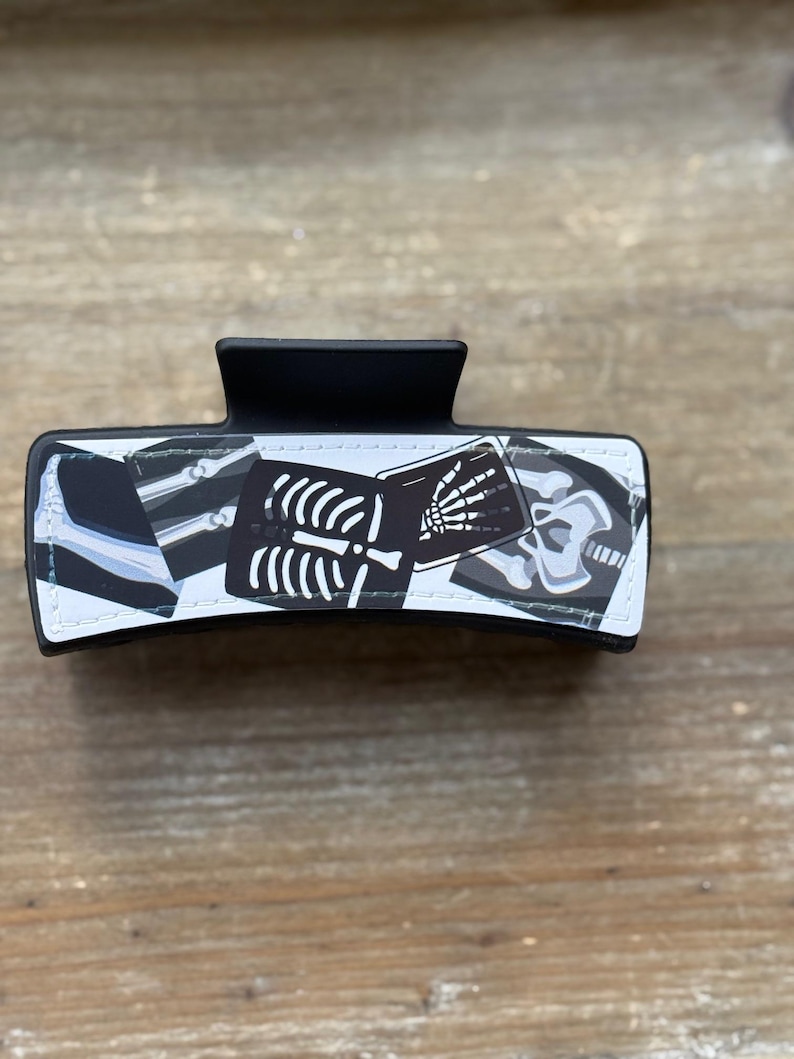 May include: Black hair claw clip featuring a design of x-ray images of bones and a skull. The clip is rectangular with a curved top and a matte finish. The x-ray images are in black and white.