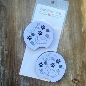 May include: Two light blue car coasters with black cat and dog paw print designs. The coasters are packaged in a clear plastic sleeve with the text "Car Coasters Handmade with love". The coasters are on a wooden surface.