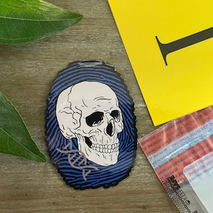 May include: A blue and white sticker with a skull and crossbones design. The skull is white with black eyes and a black outline. The crossbones are black and are in the shape of a DNA helix. The sticker is on a wooden surface.