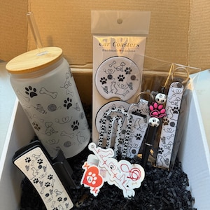 May include: A gift box filled with dog-themed items. Includes a glass tumbler with a bamboo lid and straw, car coasters, a keychain, a pen, and a car vent clip. All items feature paw prints, bones, and dog silhouettes.