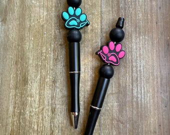 May include: Two black pens with paw print toppers. One pen has a teal paw print, and the other has a pink paw print. The pens have silver accents and are on a wooden surface.