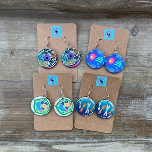 May include: Four pairs of earrings featuring different cell structures. The earrings are round and made of wood. The designs include a plant cell, an animal cell, and two pairs of chromosomes.