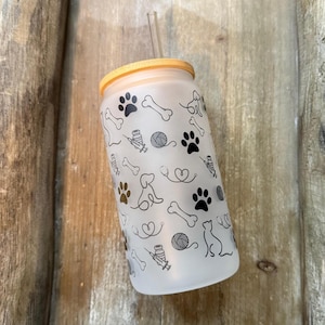 May include: A frosted glass tumbler with a bamboo lid and a clear straw. The tumbler is decorated with a repeating pattern of dog paws, bones, yarn balls, and cat silhouettes. The tumbler is sitting in a wooden tray.
