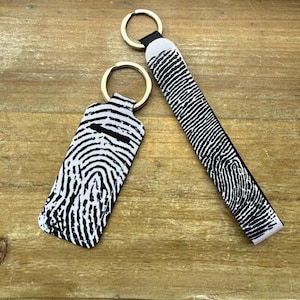 May include: Two keychains with gold-colored rings. One is rectangular, the other is a rectangle with rounded edges. Both are black and white with a fingerprint pattern. The keychains are on a wooden surface.