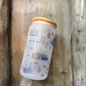 May include: A frosted glass tumbler with a bamboo lid and a clear straw. The tumbler is decorated with colorful science-themed graphics, including beakers, test tubes, and molecular structures. The design is printed on the glass.