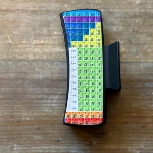 May include: A black hair claw clip featuring a colorful periodic table design. The clip has a curved shape and a matte finish. The periodic table is displayed in a rainbow of colors, with each element's symbol and atomic number visible.
