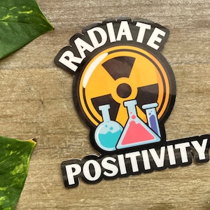 May include: Yellow and black sticker with the text "RADIATE POSITIVITY" and a stylized image of a radiation symbol with three beakers in front of it. The beakers are filled with blue, pink, and purple liquids.