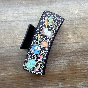 May include: A black hair claw clip featuring a repeating white cow print pattern and various colorful school supply graphics including pencils, an open book, a blue backpack, a paint palette, scissors, and a floral patterned notebook.