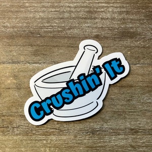 May include: A white sticker with a black outline of a mortar and pestle. The text "Crushin' It" is written in blue letters around the mortar and pestle.
