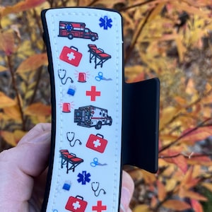 May include: A white and black silicone phone grip with a white background and a pattern of red crosses, ambulances, and medical symbols. The grip has a black clip on the side.