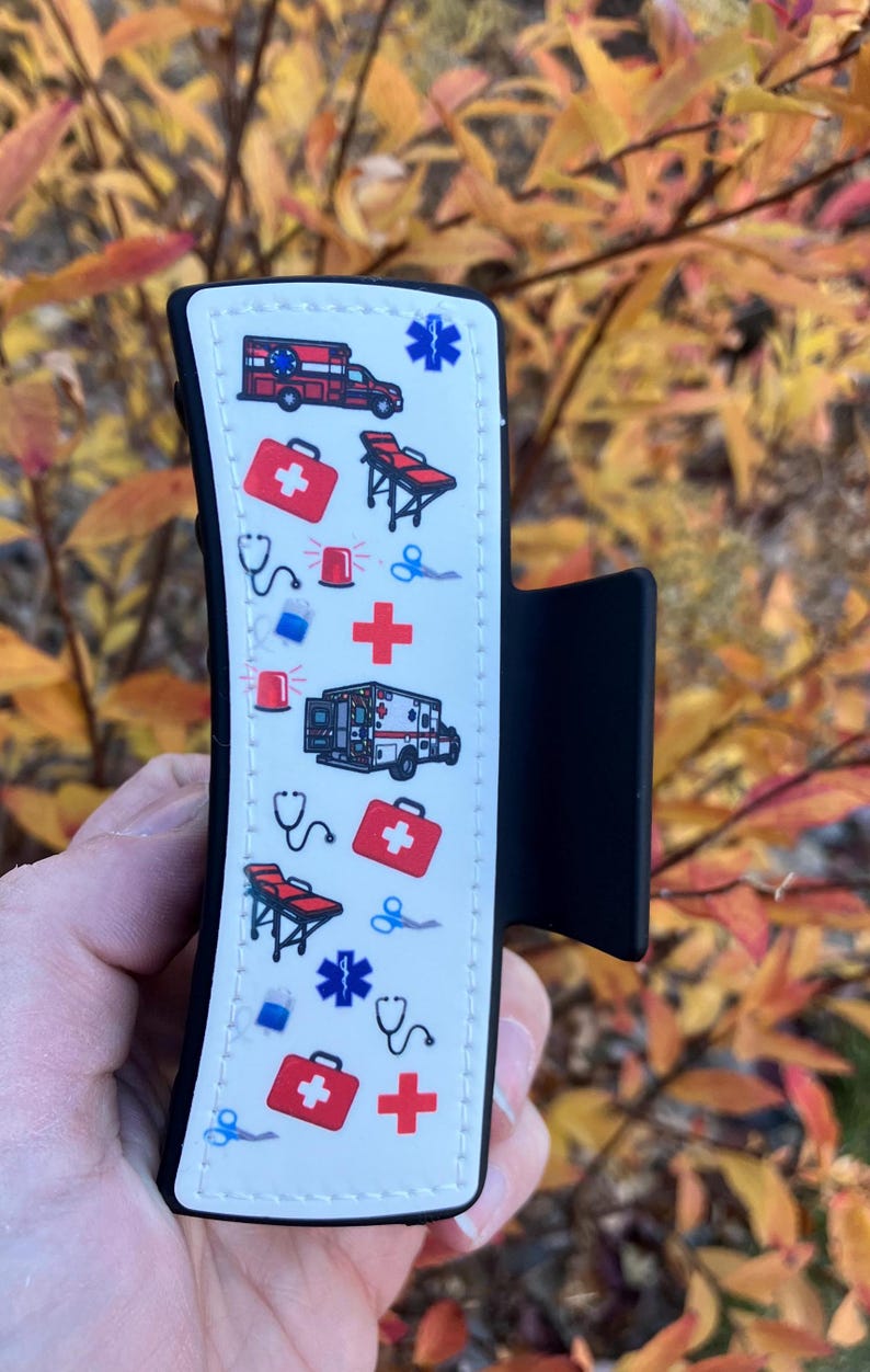 May include: A white and black silicone phone grip with a white background and a pattern of red crosses, ambulances, and medical symbols. The grip has a black clip on the side.