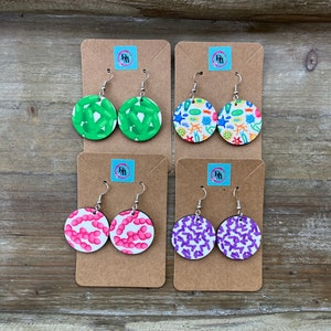 May include: Four pairs of round earrings with unique patterns. The earrings feature green, blue, pink, and purple designs. Each pair is displayed on a brown card with a logo. The earrings have silver hooks.