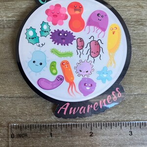 May include: A round sticker with a black border and a white background. The sticker features a variety of colorful cartoon microbes with smiling faces. The text "Cultural Awareness" is written in a pink font around the edge of the sticker.