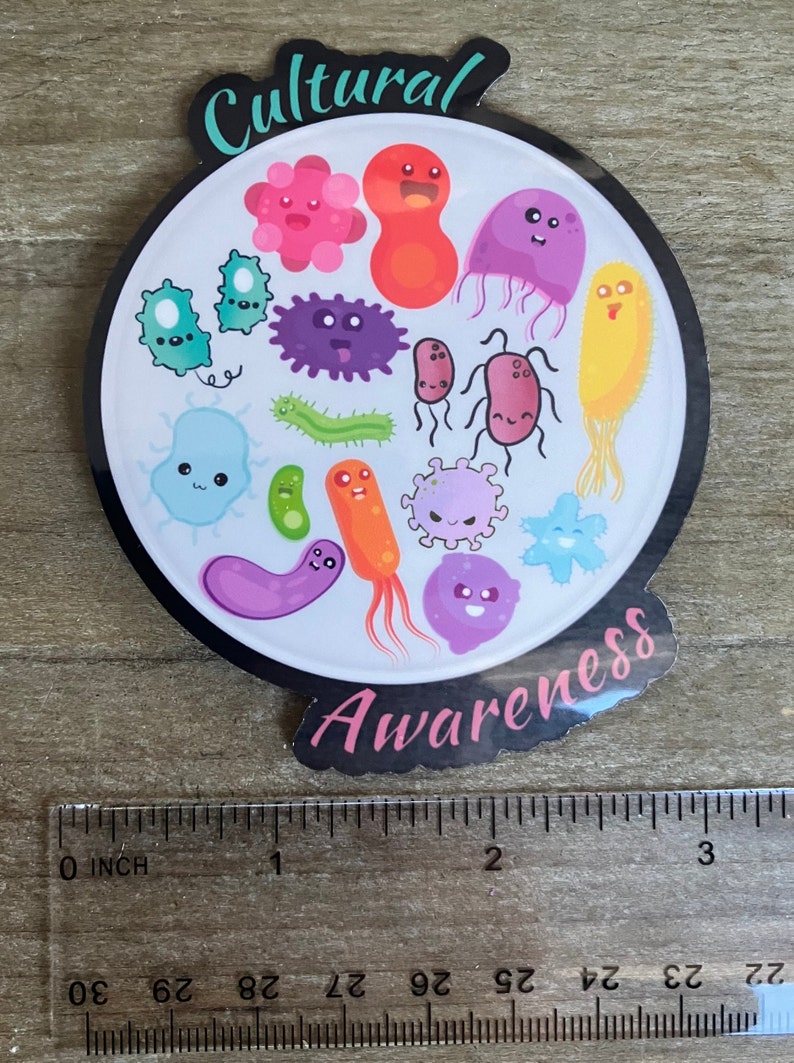 May include: A round sticker with a black border and a white background. The sticker features a variety of colorful cartoon microbes with smiling faces. The text "Cultural Awareness" is written in a pink font around the edge of the sticker.