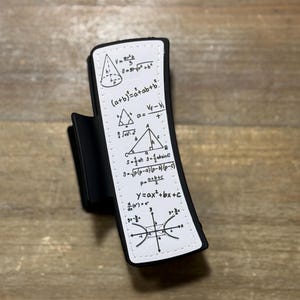 May include: Black hair clip with a white rectangular panel featuring handwritten mathematical formulas and equations. The clip is curved and has a matte finish, with a spring-loaded mechanism.