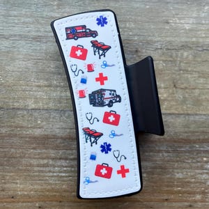 May include: A black hair clip with a white design featuring a repeating pattern of medical symbols, including ambulances, first aid kits, stethoscopes, and crosses.