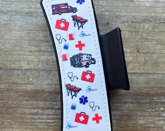 Puede incluir: Una pinza para el pelo negra con un diseño blanco que presenta un patrón repetido de símbolos médicos, incluyendo ambulancias, botiquines de primeros auxilios, estetoscopios y cruces.