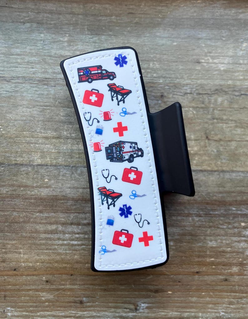 May include: A black hair clip with a white design featuring a repeating pattern of medical symbols, including ambulances, first aid kits, stethoscopes, and crosses.