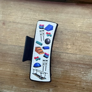 May include: A black hair claw clip featuring a white rectangular inlay decorated with a colorful pattern of physical therapy equipment, including crutches, dumbbells, a balance ball, a foam roller, a wedge, steps, and parallel bars.
