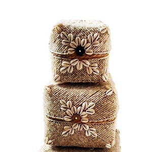May include: Three square boxes made of woven natural material, decorated with white seashells in a floral pattern. The boxes are stacked on top of each other, with the largest box on the bottom and the smallest box on top.