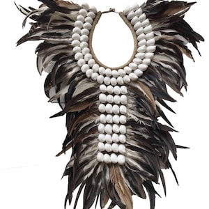 Puede incluir: Un collar de conchas blancas con un borde de plumas marrones y negras. El collar está hecho de muchas conchas pequeñas y redondas enhebradas en un cordón. Las plumas están dispuestas en forma de abanico alrededor del collar.