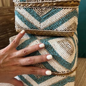 May include: Two decorative beaded boxes stacked on top of each other. The boxes are adorned with a geometric pattern of white, turquoise, and tan beads. The boxes have a woven, natural-colored border.