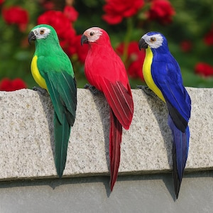 May include: Three vibrant parrot ornaments perched on a stone wall. The parrots are green, red, and blue, with yellow detailing. Red flowers are visible in the background.