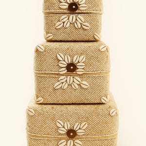 May include: A stack of four beige decorative boxes. Each box is square with a woven texture and adorned with white seashells arranged in a floral pattern. A brown button is in the center of each floral design.
