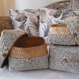 May include: A collection of woven storage baskets with lids, adorned with white beads and seashells. The baskets have a natural woven texture and are decorated with floral seashell patterns. Decorative pillows with a tropical leaf pattern are in the background.