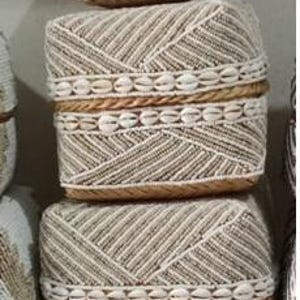Puede incluir: Tres cestas de almacenamiento rectangulares tejidas apiladas. Las cestas presentan un patrón diagonal beige y blanco, bandas de conchas marinas y un ribete de cuerda natural. Las cestas se exhiben en un estante, mostrando su diseño decorativo.