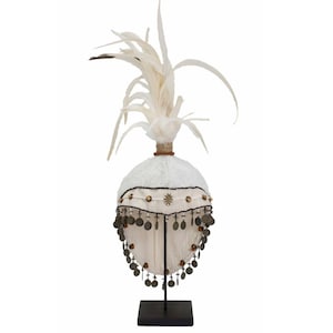 Puede incluir: Un tocado de plumas blancas con una base de tela blanca y detalles dorados y marrones. El tocado está decorado con monedas y se exhibe en un soporte negro.