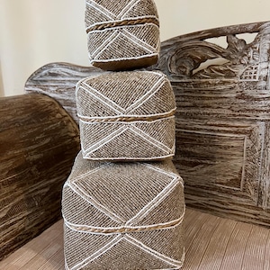 May include: Three stacked square decorative boxes. The boxes are covered in small, light brown beads with white beaded diagonal lines and a rope-like trim. The boxes are on a light brown surface with a carved wooden background.