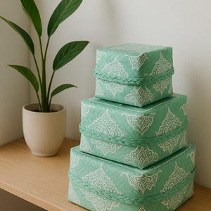 Puede incluir: Cuatro cajas de almacenamiento cuadradas apiladas, de diferentes tamaños. Las cajas son de color verde claro con un patrón blanco adornado y una textura tejida. A la izquierda hay una pequeña planta en maceta con hojas verdes.